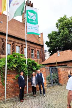 Drei Personen stehen am Fahnenmast und schauen in Richtung des Publikums, über ihnen weht die Mayors 4 Peace Flagge