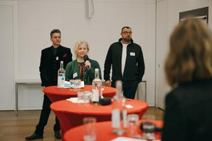 Eine Frau hält ein Mikro in der Hand und steht vor einem Tisch. Sie spricht zu einer Präsentation.