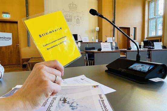 Gelbe Stimmkarte der SVV wird von einer Hand hochgehoben, im Hintergrund ein Mikro und Dokumente auf dem Tisch liegend, weitere Tische und Stühle der