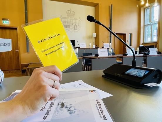 Gelbe Stimmkarte der SVV wird von einer Hand hochgehoben, im Hintergrund ein Mikro und Dokumente auf dem Tisch liegend, weitere Tische und Stühle der SVV-Sitzung und das Wappen der Stadt Brandenburg an der Havel an einer Wand
