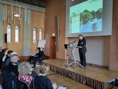 ein Mann am Rednerpult, davor sitzt Publikum und zwei Gebärdendolmetscherinnen