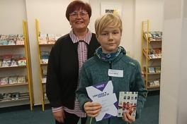 Ein Junge hält ein Buch in der Hand, hinter ihm steht die Bibliotheksleiterin.