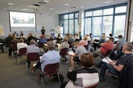 Vortragsraum mit einer Beamer-Präsentation, einer Person, die spricht, und den Zuhörerenden auf Sitzplätzen