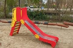 rot-gelbe Kleinkindrutsche auf dem Sandboden des Spielplatzes, im Hintergrund das Schlangenspielgerät