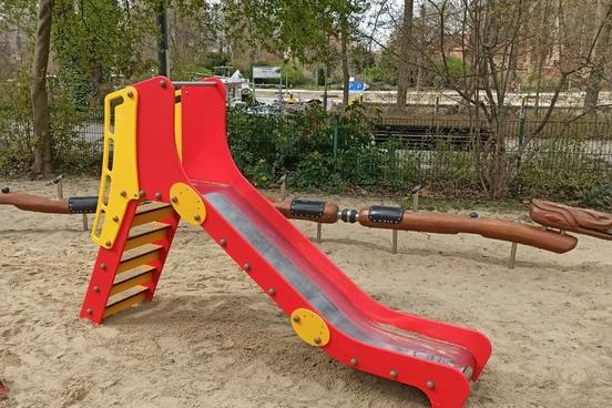 rot-gelbe Kleinkindrutsche auf dem Sandboden des Spielplatzes, im Hintergrund das Schlangenspielgerät
