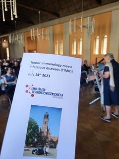 Flyer der Veranstaltung im Vordergrund, dahinter eine Sprecherin am Pult und das Publikum im Rolandsaal