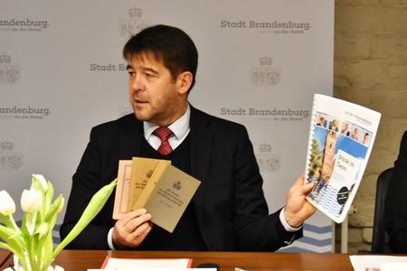 OB Steffen Scheller hält alte, kleine,  braune Hefte und ein A4 großes bunt ausgedrucktes Dokument hoch