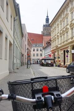 im Vordergrund ein Fahrradlenker mit Korb, im Hintergrund eine Gasse der Brandenburger Neustadt und der Kirchturm der St. Katharinenkirche