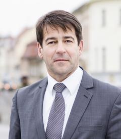 Portraitfoto von Steffen Scheller im grauen Anzug, im Hintergrund verschwommen die Jahrtausendbrücke