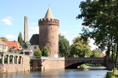 Turm neben einer Brücke direkt am Wasser, daneben weitere historische Bauten und Bäume