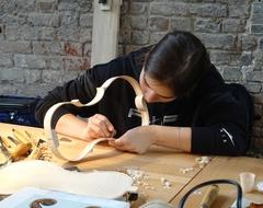 Eine Frau fertigt eine Geige, der Rohkörper der Geige liegt vor ihr auf einem Tisch.