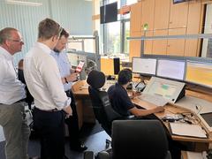 3 Männer stehen hinter einem Tisch eines Leitstellen-Disponenten. Die stehenden Männer schauen auf ein Handy mit der App nora, der Disponent schaut auf dem Bildschirm auf die Anwendung nora.