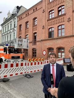 Der Oberbürgermeister spricht mit einer Journalistin, im Hintergrund eine Feuerwehr vor einem Gebäude