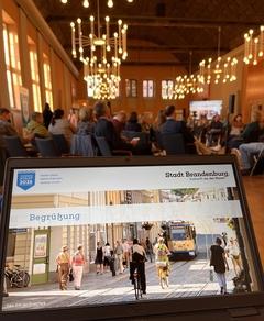 Im Hintergrund sieht man den beleuchteten Rolandsaal mit vielen Gästen. Im Vordergrund ist der Bildschirm eines Tabletts mit der Begrüßungsfolie zu se