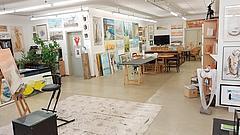 Bild zeigt das Atelier von der Künstlerin Regina Heinich, bunt und vollgestellt mit Bildern der Künstlerin. In der Mitte ein großer Arbeitstisch.