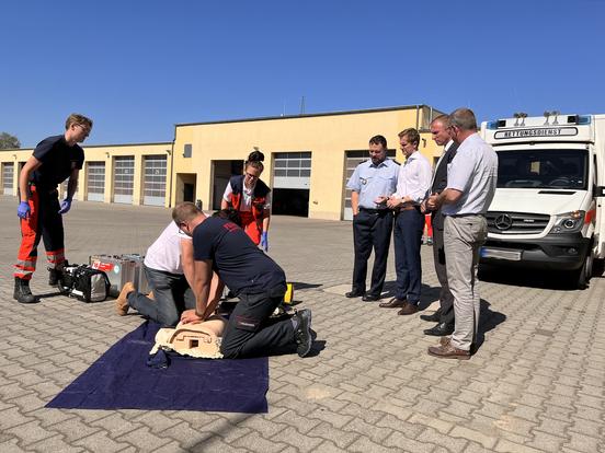 vier Männer stehen vor einem Rettungswagen, daneben wird von zwei Laien und zwei Rettungssanitätern eine Herz-Druck-Massage an einer Übungspuppe durchgeführt
