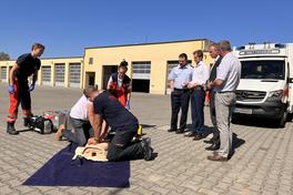 vier Männer stehen vor einem Rettungswagen, daneben wird von zwei Laien und zwei Rettungssanitätern eine Herz-Druck-Massage an einer Übungspuppe durchgeführt