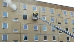 Drehleiter der Feuerwehr mit Feuerwehrleuten im Rettungskorb am Fenster eines Wohnblockes, aus dem Rauch aufsteigt
