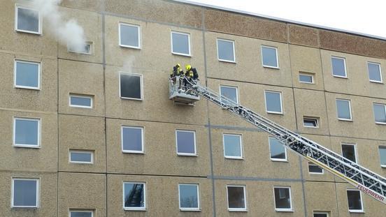 Drehleiter der Feuerwehr mit Feuerwehrleuten im Rettungskorb am Fenster eines Wohnblockes, aus dem Rauch aufsteigt