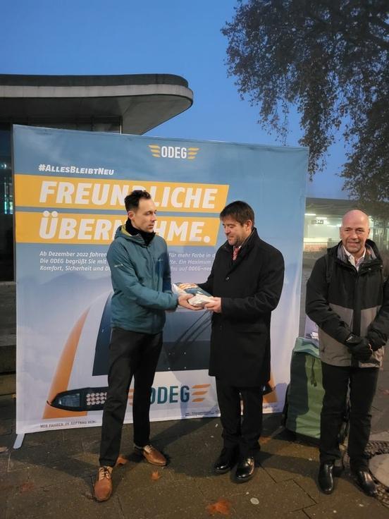 Oberbürgermeister Steffen Scheller nimmt im Beisein von Thomas Dill, Bereichsleiter des Verkehrsverbundes Berlin-Brandenburg (r.), aus den Händen von ODEG-Geschäftsführer Roland Pauli (l.) das "EinZugBrot" entgegen.