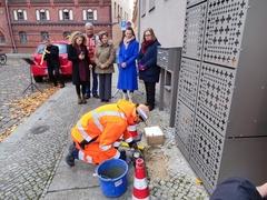ein Mensch in Arbeitskleidung hockt auf dem Kopfsteinpflasterboden mit Werkzeug; dahinter steht eine Gruppe Menschen und schaut zu