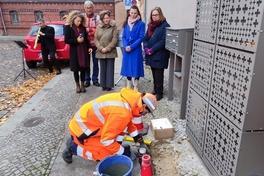 ein Mensch in Arbeitskleidung hockt auf dem Kopfsteinpflasterboden mit Werkzeug; dahinter steht eine Gruppe Menschen und schaut zu