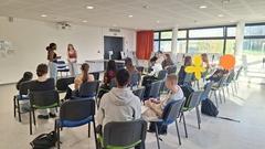 viele Schüler sitzen in einem Raum mit Glasfront. Zwei Schüler stehen vorn
