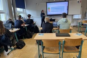 Neben dem Smart-Board steht eine Frau und hält eine Präsentation. Vor Ihr sitzen Schülerinnen und Schüler.