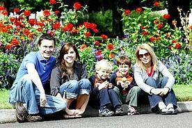 Familie sitzt auf einer Bordsteinkante und lächelt, im Hintergrund blühende Blumen