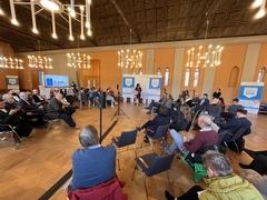 Viele Gäste sitzen in einer Art Stuhlkreis um das Rednerpult herum und beobachten eine Frau die davor steht und redet.