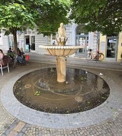 Angler-Statue auf einem Brunnen der von Bäumen umgeben ist