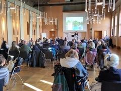 viele Menschen sitzen in einem Saal und hören Oberbürgermeister Scheller zu, der die Menschen zur Veranstaltung begrüßt.