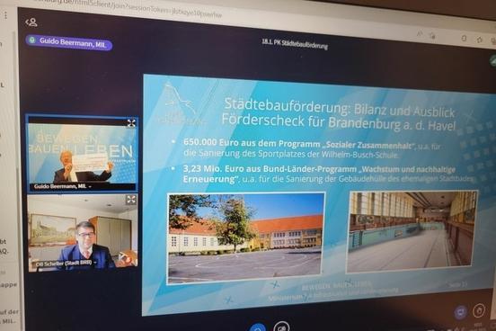 Monitor, auf der eine Videokonferenz von Guido Beermann und Steffen Scheller und eine Präsentationsfolie zu sehen ist.