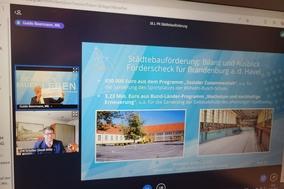 Monitor, auf der eine Videokonferenz von Guido Beermann und Steffen Scheller und eine Präsentationsfolie zu sehen ist.