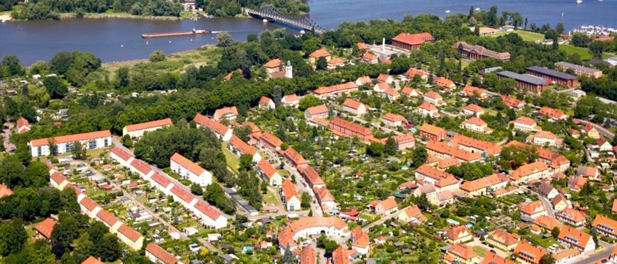 Luftbild mit vielen roten Dächern, Grünflächen und einem See im Hintergrund