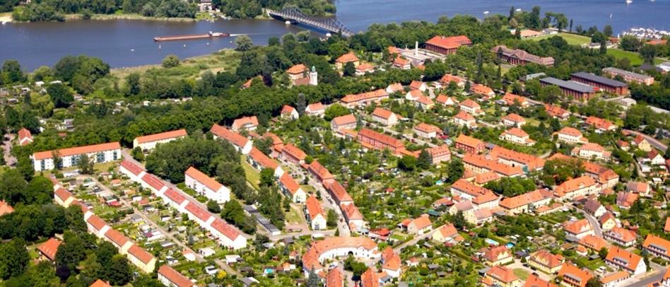 Luftbild mit vielen roten Dächern, Grünflächen und einem See im Hintergrund