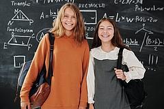 Zwei Mädchen mit Schultasche vor einer Tafel