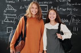 Zwei Mädchen mit Schultasche vor einer Tafel