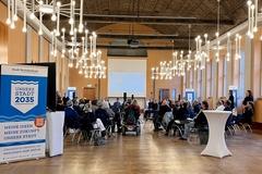 Viele Menschen sitzen in einer Art Stuhlkreis im Rolandsaal. Der Saal ist beluchtet und im Vordergrund steht ein blaues Roll-Up zum Leitbild.